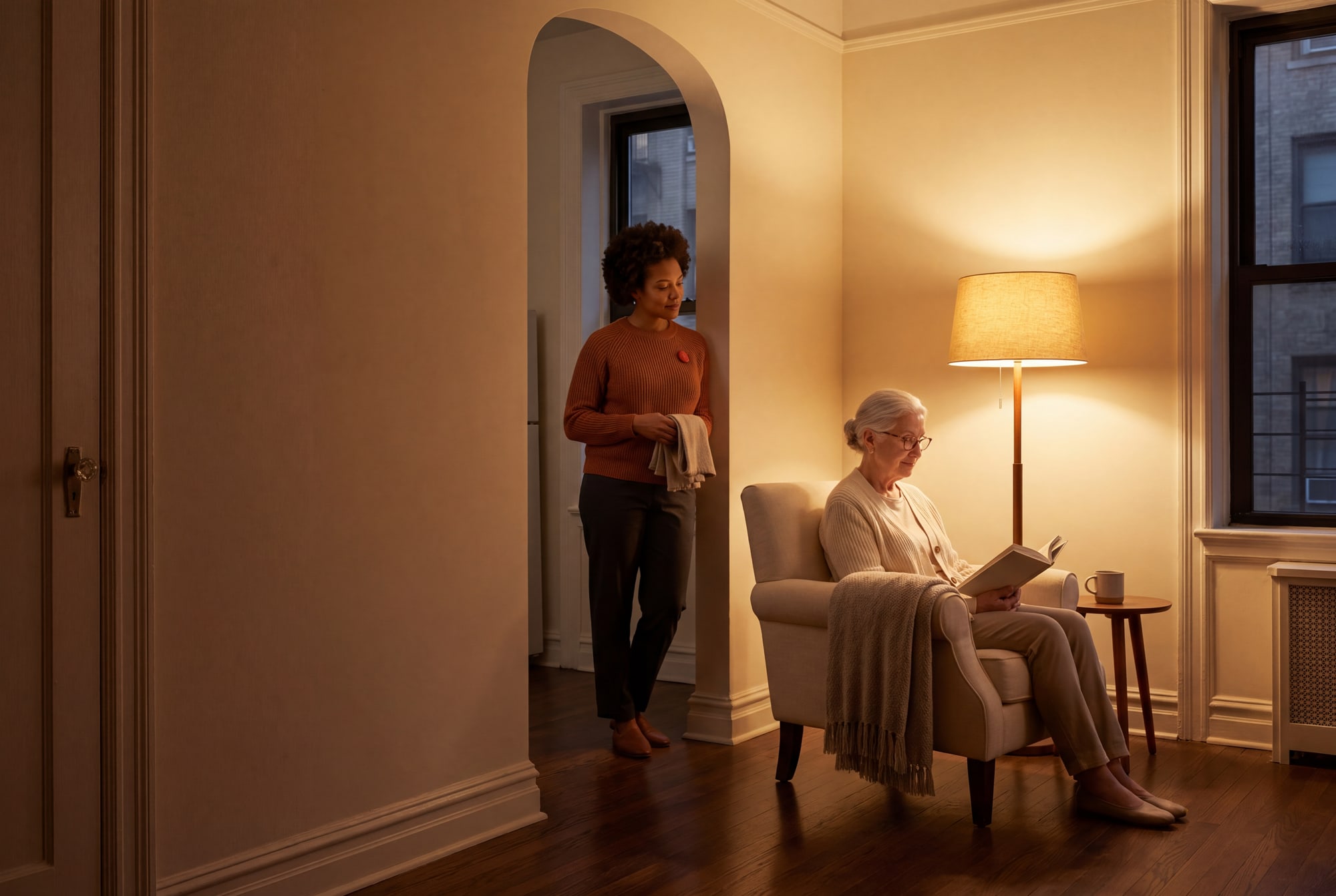 A warmly lit living room at evening. An older woman reads in an armchair under a floor lamp.
