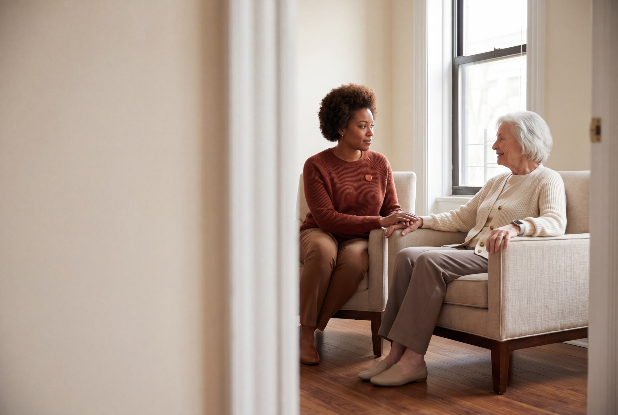An older woman sitting quietly in soft daylight, her hand resting on a familiar surface.